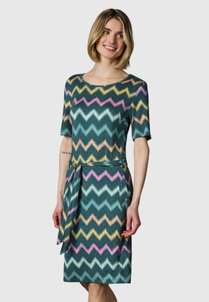Kvinde iført en knælængde kjole med flerfarvede zigzag mønstre og korte ærmer, smilende med hænder afslappet langs siderne.