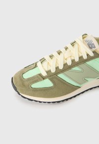 Sneaker New Balance vert et beige avec des lacets crème, des panneaux en daim et tissu, et une semelle en caoutchouc noir sur fond blanc.