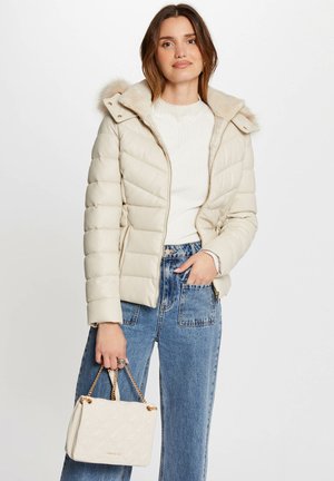 Giacca a vento beige chiaro con cappuccio in pelliccia sintetica, presenta un design trapuntato e una zip dorata. Abbinata a jeans a vita alta e a una borsa leggera.