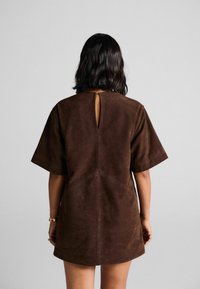 Robe en daim marron avec des manches courtes, un détail en forme de clé à l'arrière et une coupe droite. Texture lisse avec des accents de couture subtils.