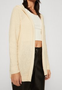 Žena v krémovém otevřeném pleteném kardiganu přes bílý žebrovaný crop top a černé kalhoty vysokého pasu, stojící před čistým pozadím.