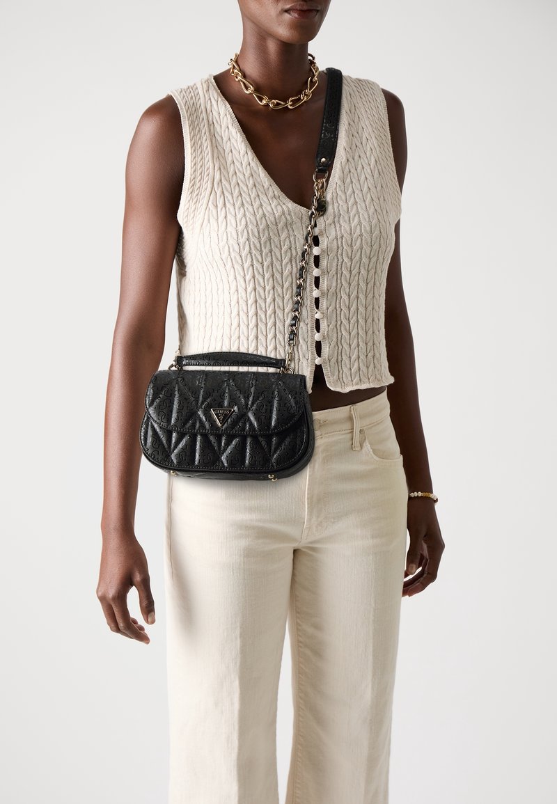 Femme portant un gilet beige en maille sans manches, un pantalon crème, un collier chaîne doré, un bracelet et un sac bandoulière noir matelassé avec une bandoulière chaîne.