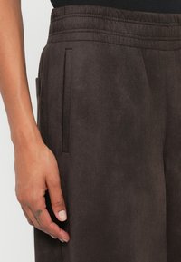 Pantaloni din suede maro cu talie elastică, prevăzuți cu buzunare laterale și o textură moale, prezentați cu o mână poziționată aproape de buzunar.