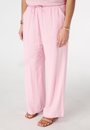 Vrouw draagt losse, lichtroze broek met trekkoord in de taille, gecombineerd met bruine sandalen met open tenen en gouden armbanden om beide polsen.