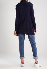 Marinblå långärmad cardigan med en lös passform, lätt material och sidofickor, kombinerad med uppkavlade jeans i denim och vita sneakers.