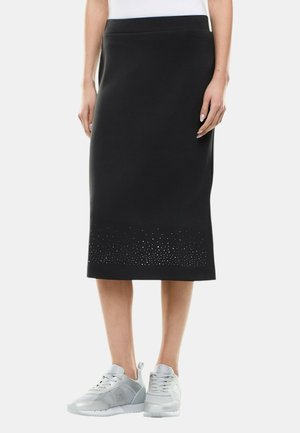 Donna che indossa una gonna a matita nera al ginocchio con piccole decorazioni vicino all'orlo e sneakers grigio chiaro.