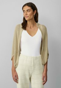 Beige, gestrickte Cardigan mit offener Vorderseite und weiten Ärmeln, kombiniert mit einem weißen V-Ausschnitt-Bodysuit und strukturierten cremefarbenen Hosen.