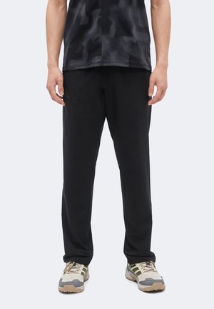 Homme portant un pantalon droit noir, une chemise noire à motifs et des baskets gris-beige avec des accents verts, debout devant un fond uni.