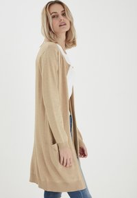 Gilet long beige en tricot doux et texturé. Peut être porté ouvert, avec des poignets côtelés et deux poches avant. Associé à un haut blanc et un jean.