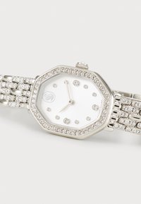 Silberne achteckige Uhr mit weißem Ziffernblatt, verziert mit Kristallakzenten. Metallarmband mit miteinander verbundenen Segmenten und funkelnden Steinen.
