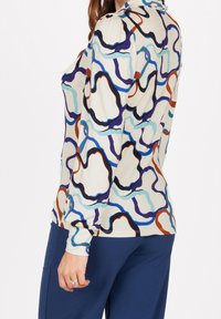 Pitkähiha toppi vaalean kermanvärisestä kankaasta, jossa on värikkäitä abstrakteja käärmeennäköisiä kuvioita sinisessä, mustassa, punaisessa ja violetissa; pehmeä tekstuuri, vartalonmyötäinen muotoilu.