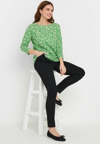 Grüne Blumenbluse mit Dreiviertelärmeln, kombiniert mit schwarzen Skinny-Jeans und schwarzen Ballerinas mit einer Schleifenapplikation.