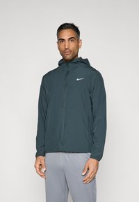 Tumši zaļzaļa viegla kapuce jakas ar priekšā rāvējslēdzēju un elastīgiem aprocēm, uzlabota ar mazu balto Nike logotipu kreisajā krūtīs.