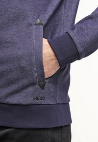 Felpa blu navy con tessuto testurizzato, dotata di una tasca nascosta, dettagli a triangolo in pelle e polsini a costine. Logo del marchio visibile all'orlo.
