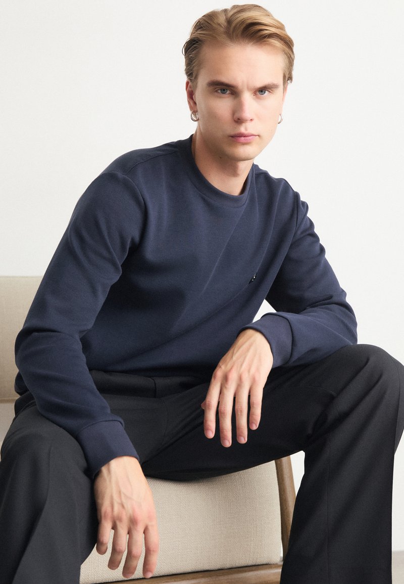 Marinblå sweatshirt i mjukt tyg med rund halsringning, ribbade ärmslut och en subtil logotypdetalj på bröstet. Styler med svarta byxor.