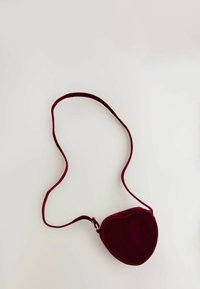 Torebka crossbody w kształcie serca wykonana z burgundowego zamszu, z cienkim paskiem i widocznymi detalami szwów. Gładka tekstura o minimalistycznym designie.
