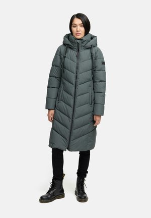 Longo cappotto imbottito con cappuccio di colore verde scuro, caratterizzato da un design a chevron, zip frontale, cordini regolabili e tasche laterali. Indossato con pantaloni neri e stivali.