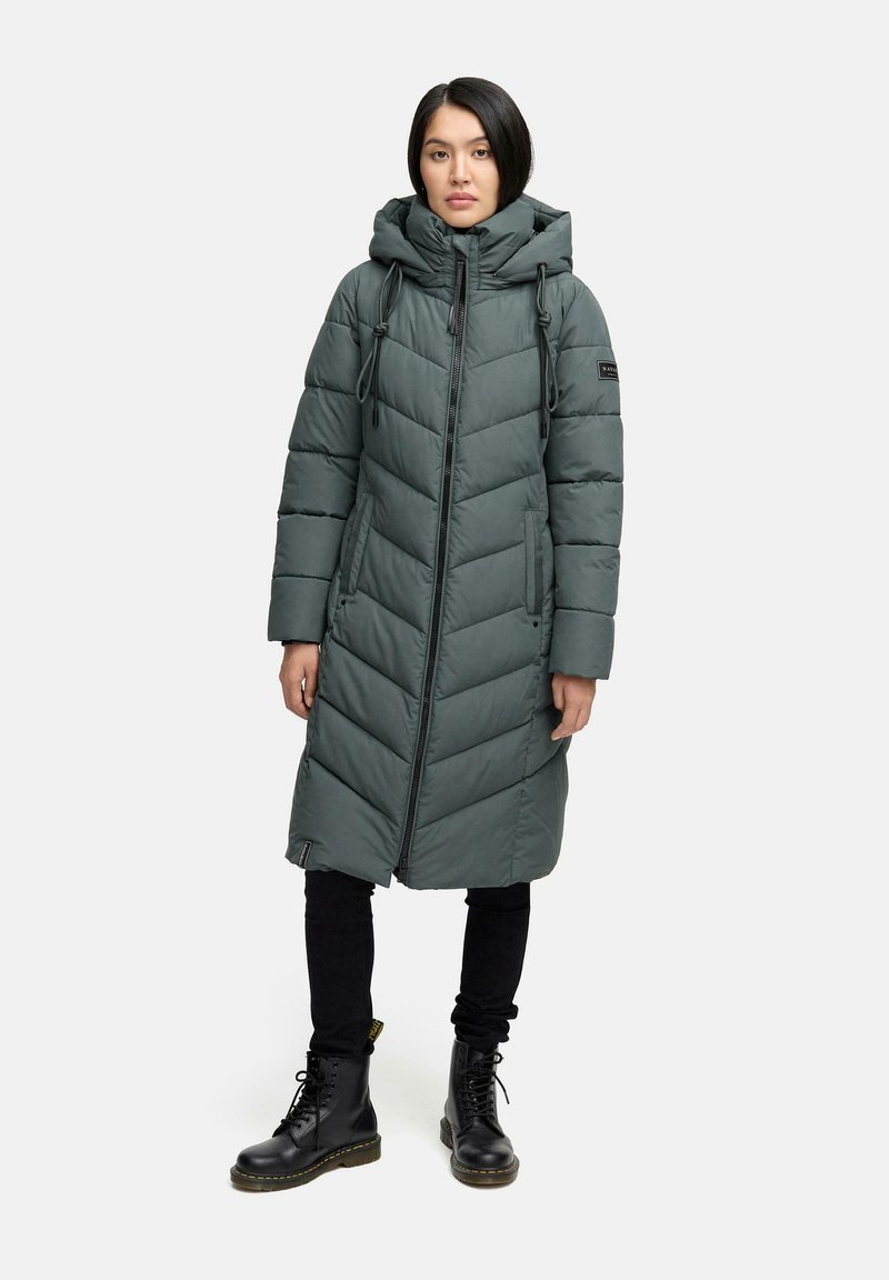 Lange, gebreide pufferjas met capuchon in donkergroen, met een chevrondesign, een rits aan de voorkant, verstelbare trekkoorden en zijzakken. Gecombineerd met zwarte broek en laarzen.