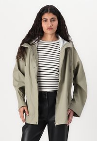 Veste imperméable vert olive avec capuche, dotée d'une fermeture éclair à double sens, de boutons-pression externes et d'une doublure intérieure blanche, portée sur un haut rayé.