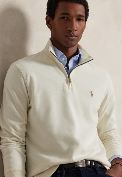 Άνδρας που φοράει εκρού πουλόβερ με φερμουάρ μέχρι το στήθος πάνω από ανοιχτό μπλε πουκάμισο με γιακά, σκούρο τζιν και δερμάτινη ζώνη πάνω σε ουδέτερο φόντο.