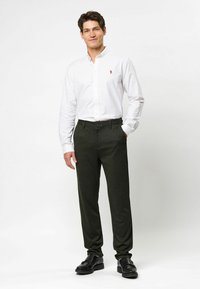 Balta mygtukinė marškiniai, žali kelnės ir juodi batai. Marškiniai turi mažą siuvinėtą logotipą; kelnės yra pritaikytos, su lygiu paviršiumi.