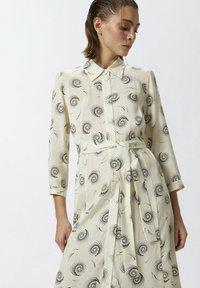 Abito camicia crema con un motivo nero a spirale, maniche lunghe, colletto e dettaglio in vita annodato. Il tessuto appare liscio e leggero.