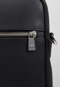 Sac en cuir texturé noir avec une fermeture éclair argentée et un tire-zip métallique présentant un petit logo gravé et un détail à rayure bleue.