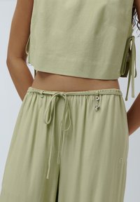 Top corto verde chiaro con lacci sui lati, abbinato a pantaloni larghi coordinati con vita elasticizzata e un charm decorativo.