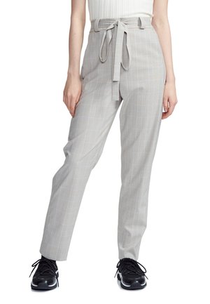 Kobieta ubrana w jasnoszare, wysokie spodnie w kratę z wiązanym paskiem, biały top bez rękawów oraz czarne sneakersy z białymi podeszwami.