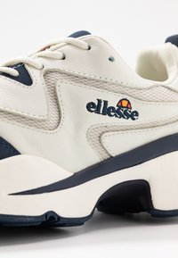 Scarpa sportiva bianca con pannelli in mesh e pelle, dettagli blu navy e lacci. Presenta il logo "ellesse" ricamato di lato. Suola in gomma piatta.