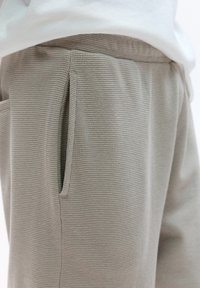 Sive tkanine joggerice s teksturom, koje sadrže bočne džepove i elastičnu pojas, prikazane iz neposredne blizine s naglaskom na materijal i detalje.