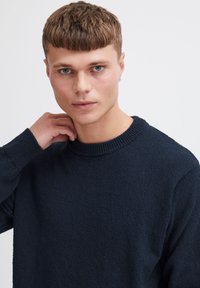 Maglione lavorato a maglia di colore blu navy con collo rotondo, tessuto testurizzato e polsini a coste. Il modello si tocca il collo, evidenziando un fit rilassato e morbidezza.
