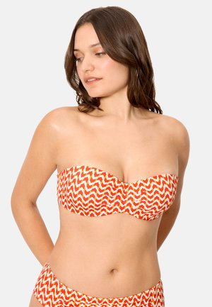 Mujer con bikini sin tirantes con patrón de zigzag en naranja y blanco, mirando hacia abajo y hacia un lado contra un fondo liso.