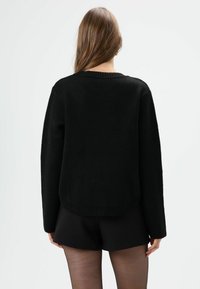 Schwarzer Strickpullover mit rundem Ausschnitt, überschnittenen Schultern und geripptem Saum. Die Textur wirkt weich und leicht dehnbar. Kombiniert mit schwarzen Shorts.