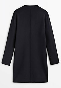 Manteau en tricot noir avec manches longues, col montant et coupe droite. Présente une couture centrale à l'arrière et des poignets côtelés.