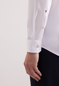 Camicia bianca a maniche lunghe con polsino arrotondato, caratterizzata da un singolo bottone dorato e una piccola rosa nera ricamata sulla manica.