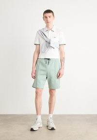 Polo blanc à manches courtes avec des accents verts, associé à un short vert menthe. Un pull gris clair est drapé sur les épaules. Baskets blanches.