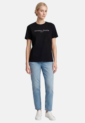 Mujer con el pelo rubio recogido en un moño, lleva una camiseta negra de Maison Gaelle, vaqueros azul claro y zapatillas blancas, está de pie frente a un fondo liso.