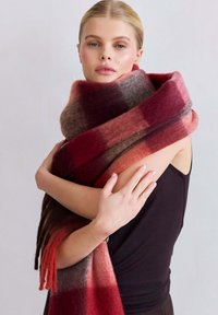 Écharpe en laine présentant de larges rayures en dégradé de rouge, marron et gris. Texture douce avec des extrémités à franges, enroulée autour des épaules d'un modèle.