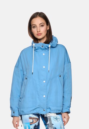 Frau trägt eine hellblaue Kapuzenjacke mit weißen Kordeln und bedruckte Leggings mit nautischen Motiven.