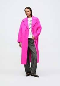 Sieviete, tērpusies spilgti rozā garā mētelī virs baltas apdrukātas krekla un melnām platām biksēm, stāv ar vienu roku kabatā.