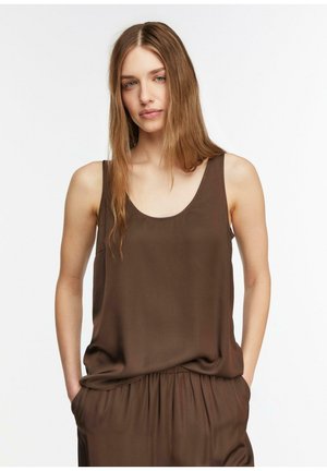 Femme aux longs cheveux châtain clair portant un haut marron sans manches et un pantalon ample assorti, debout les mains dans les poches sur fond blanc.