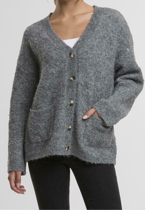 Kvinde iført en blød grå cardigan med knapper foran og lommer, kombineret med sorte bukser og en hvid undershirt synlig ved halsudskæringen.
