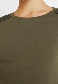 Sudadera verde oliva con cuello redondo, textura acanalada y tachuelas negras decorativas en las costuras de los hombros. Tejido suave, diseño informal.