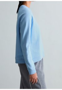 Personne portant un sweat-shirt bleu clair et un pantalon de jogging gris se tient de profil devant un fond uni gris clair.