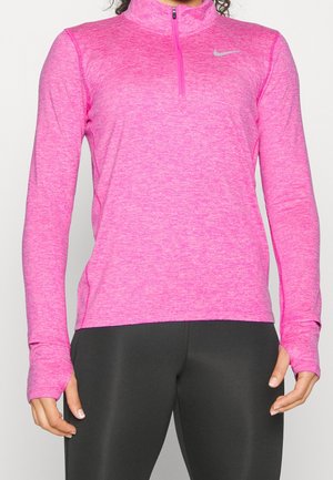 Femme portant un haut de sport rose vif à manches longues avec des trous pour les pouces et une fermeture éclair quart de tour, associé à des leggings noirs ajustés.