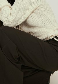 Dicker cremefarbener Strickpullover, der über dunklen, lockeren Stoffhosen getragen wird. Der Pullover hat eine gerippte Textur und ein gerafftes Bindedetail am Saum.