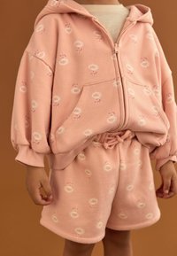 Rosa bomullströja med blommönster, dragkedja fram och sidofickor, tillsammans med matchande shorts med samma mönster och dragsnöre i midjan.
