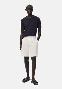 Marineblaues T-Shirt mit kurzen Ärmeln kombiniert mit weißen, gestreiften Shorts. Das Model trägt schwarze Sandalen. Einfaches, zeitgemäßes Design mit lockerer Passform.