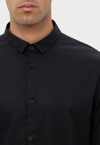 Zwarte button-up shirt met een puntige kraag, voorzien van een subtiele geborduurde logo op de borst, gladde stof en standaard knoopsluiting.